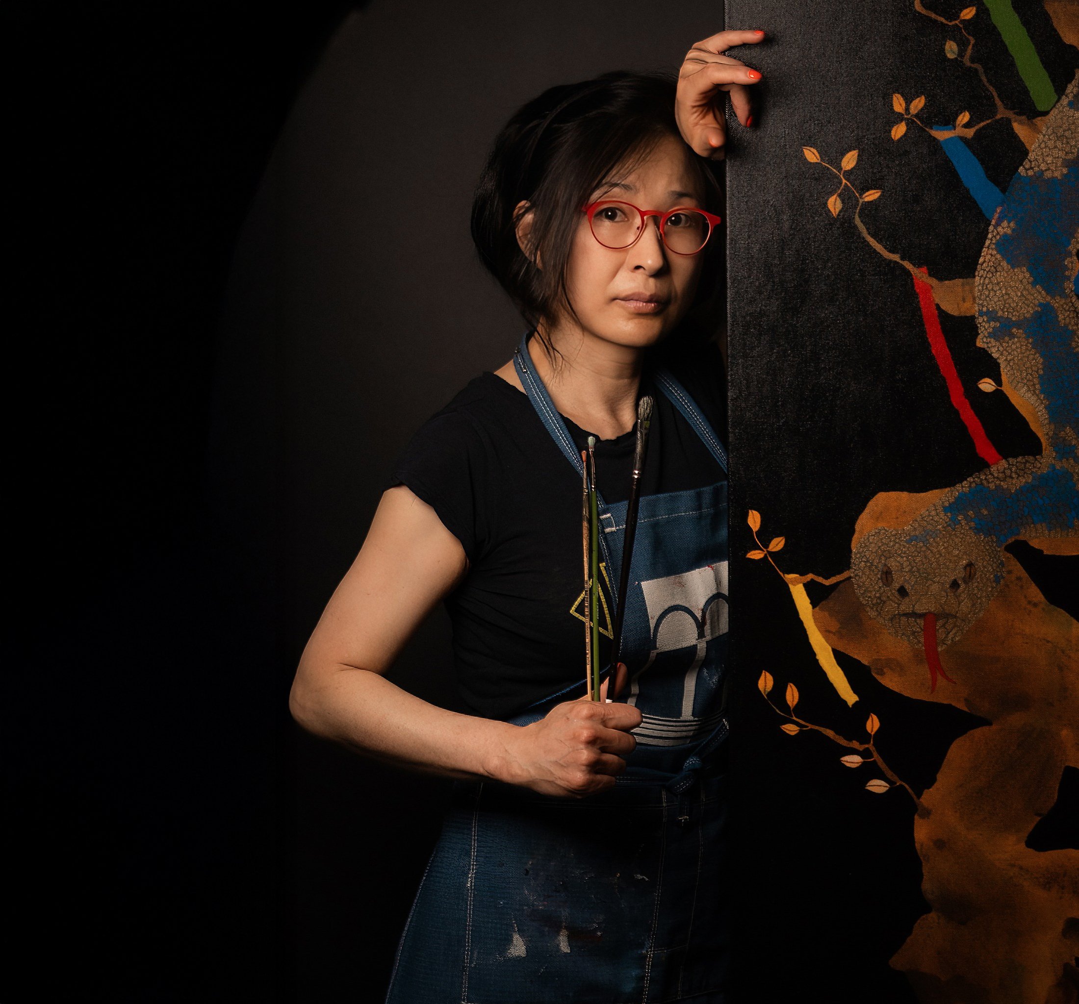 Artist with red glasses and paintbrushes beside a painting